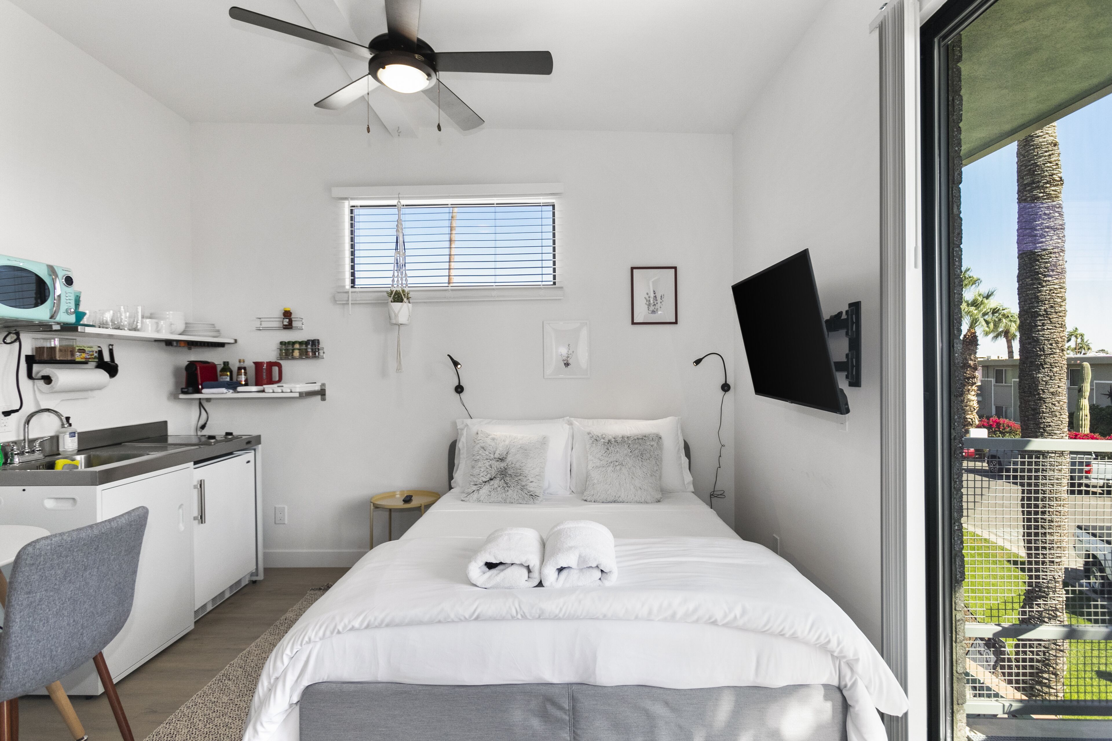A modern hotel room with a bed, a desk, a chair, a TV, and a window with blinds.