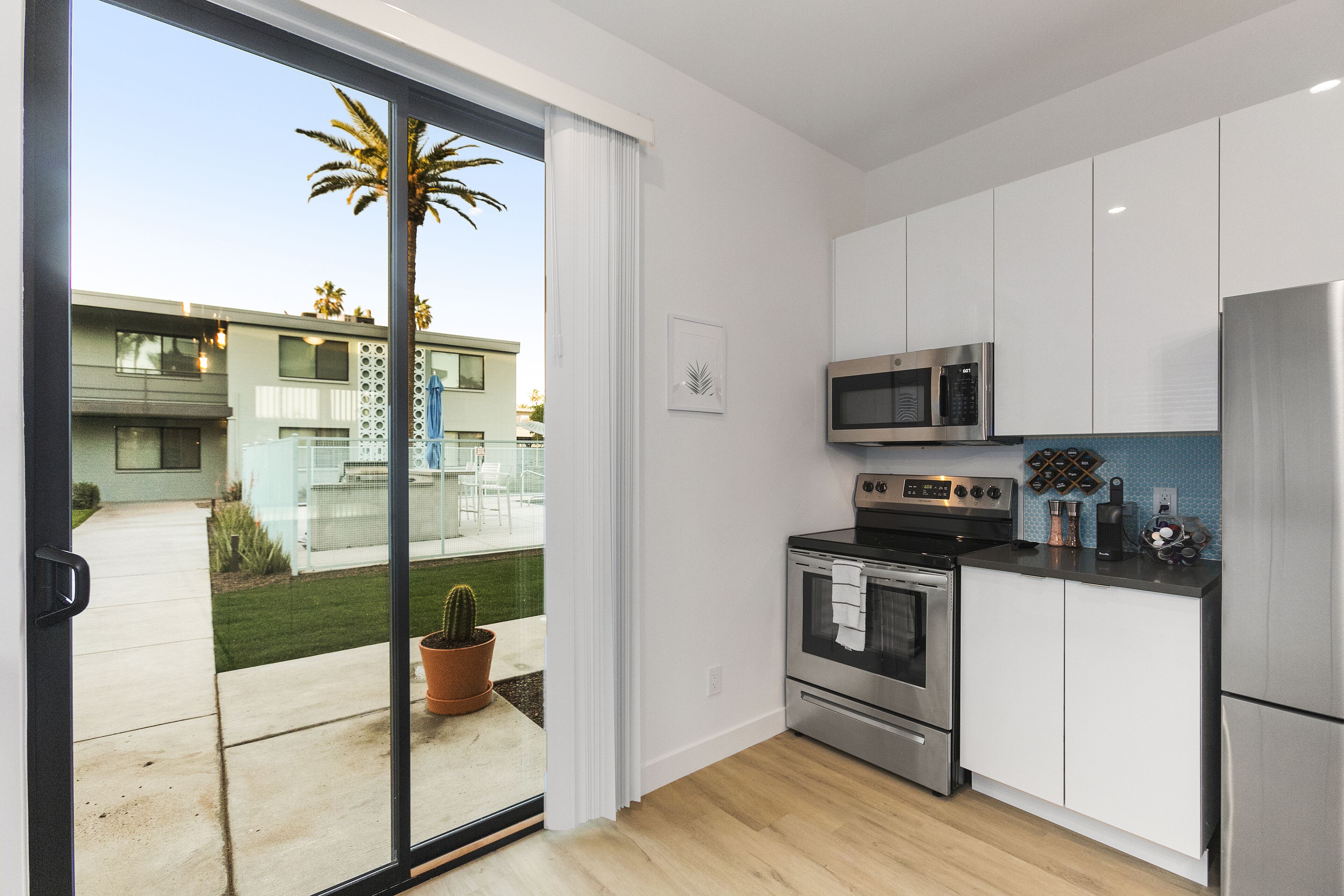 Una cuina moderna amb armaris blancs, electrodomèstics d'acer inoxidable i una porta corredissa de vidre que dona a un patí amb un cactus i vistes a edificis veïnals.