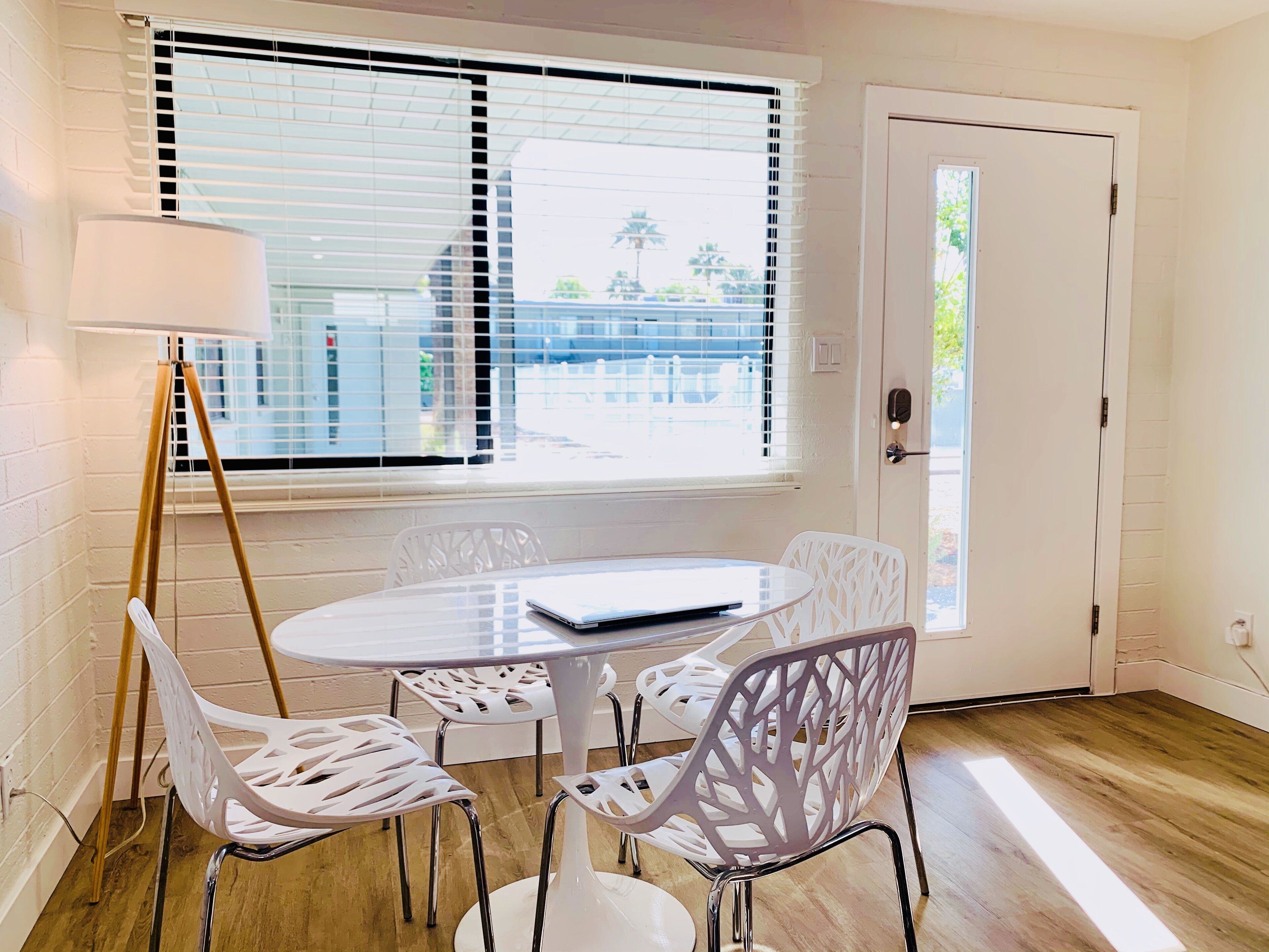 Une pièce avec une table ronde, quatre chaises blanches, un lampadaire et une fenêtre avec des stores.