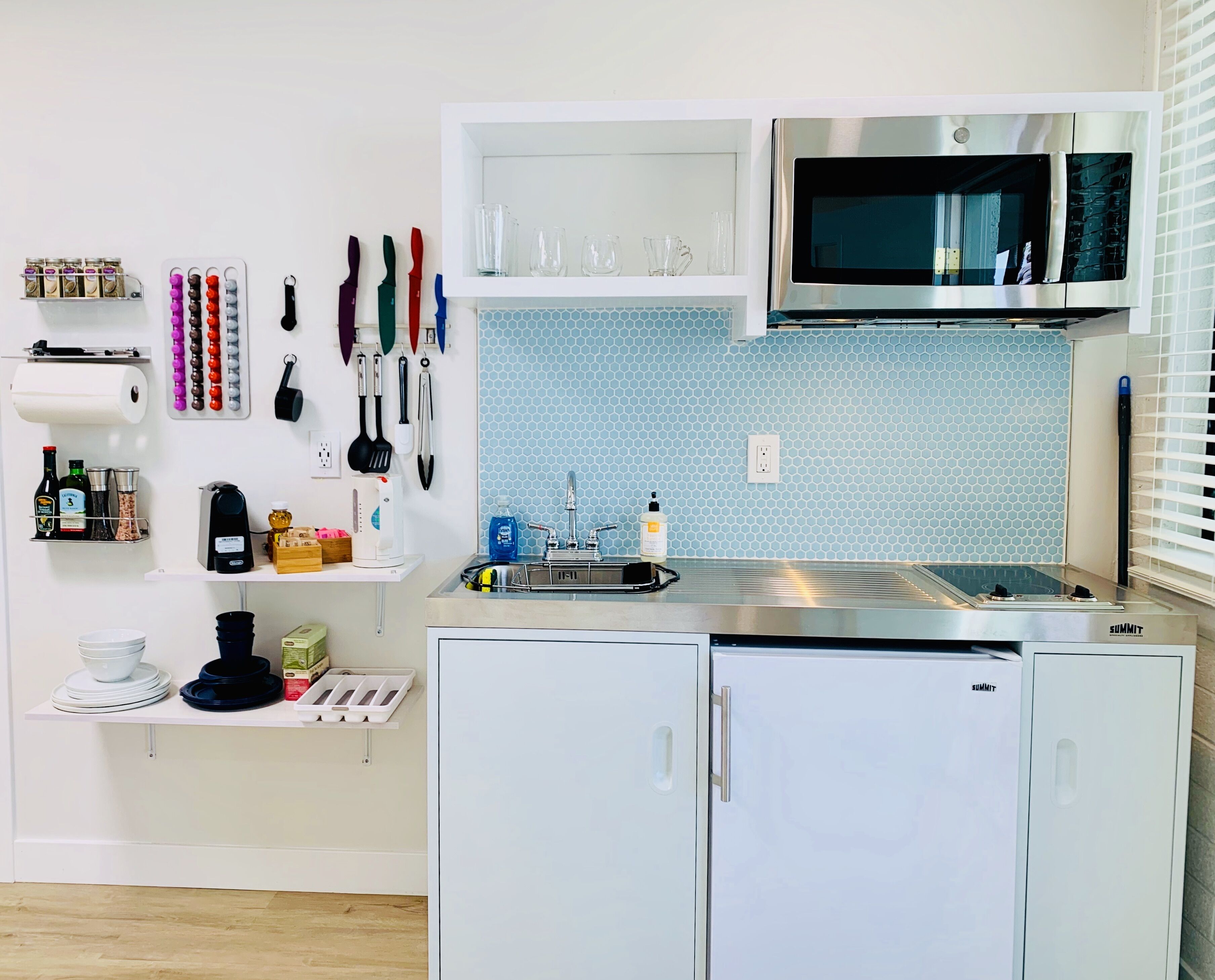 A compact kitchen with a microwave, sink, and two cabinets. There are various kitchen utensils and appliances on the wall, including a coffee maker and a spice rack.