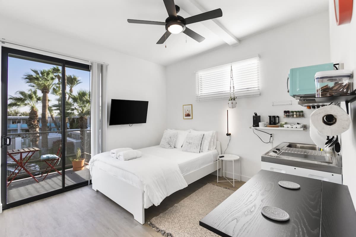 A modern bedroom with a bed, a ceiling fan, a TV, a small kitchen area with a sink, and a balcony with a chair and potted plant.