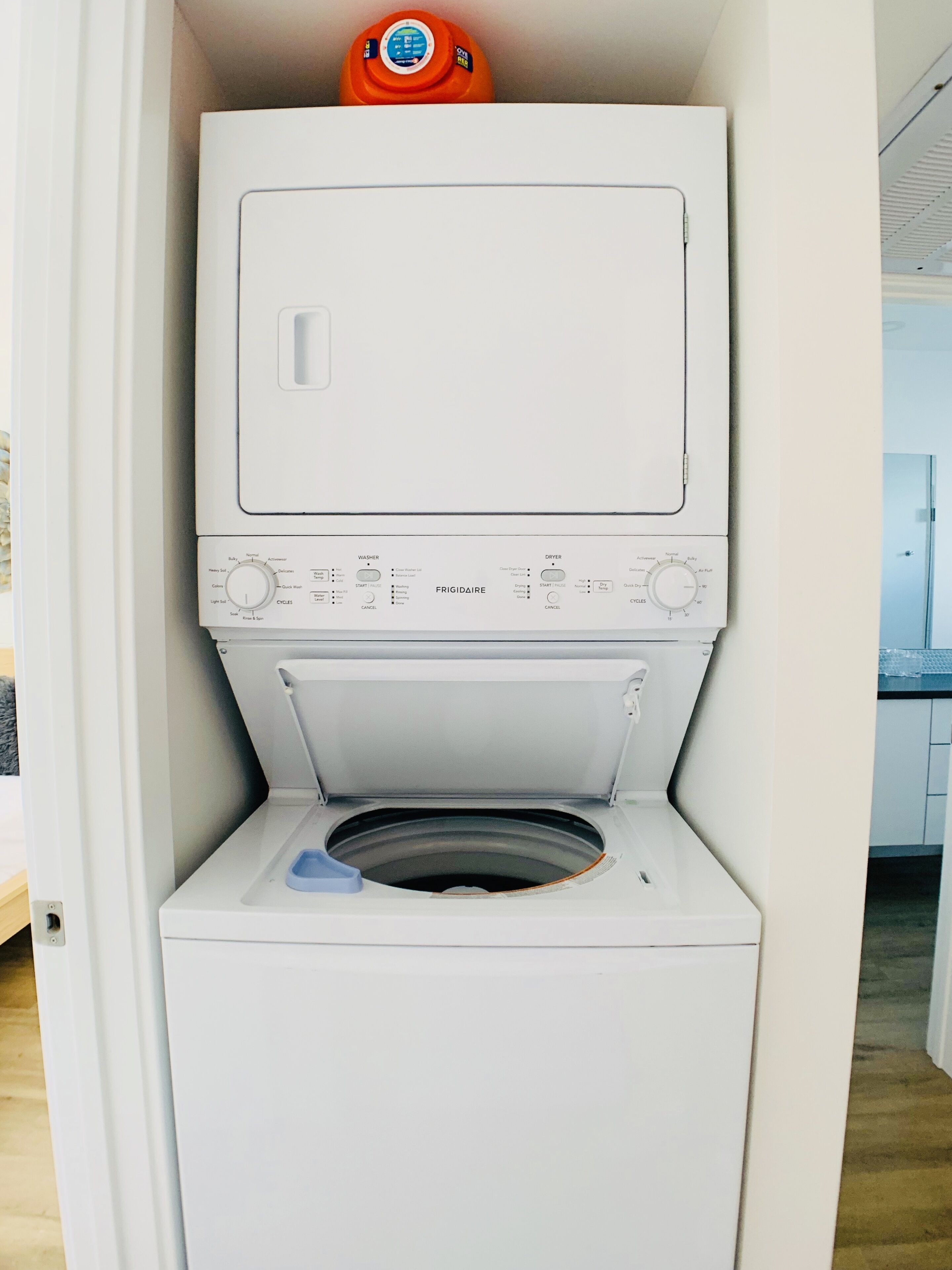 Lavadora blanca compacta con puerta frontal, panel de control con varias opciones y un pequeño tanque de agua naranja en la parte superior.