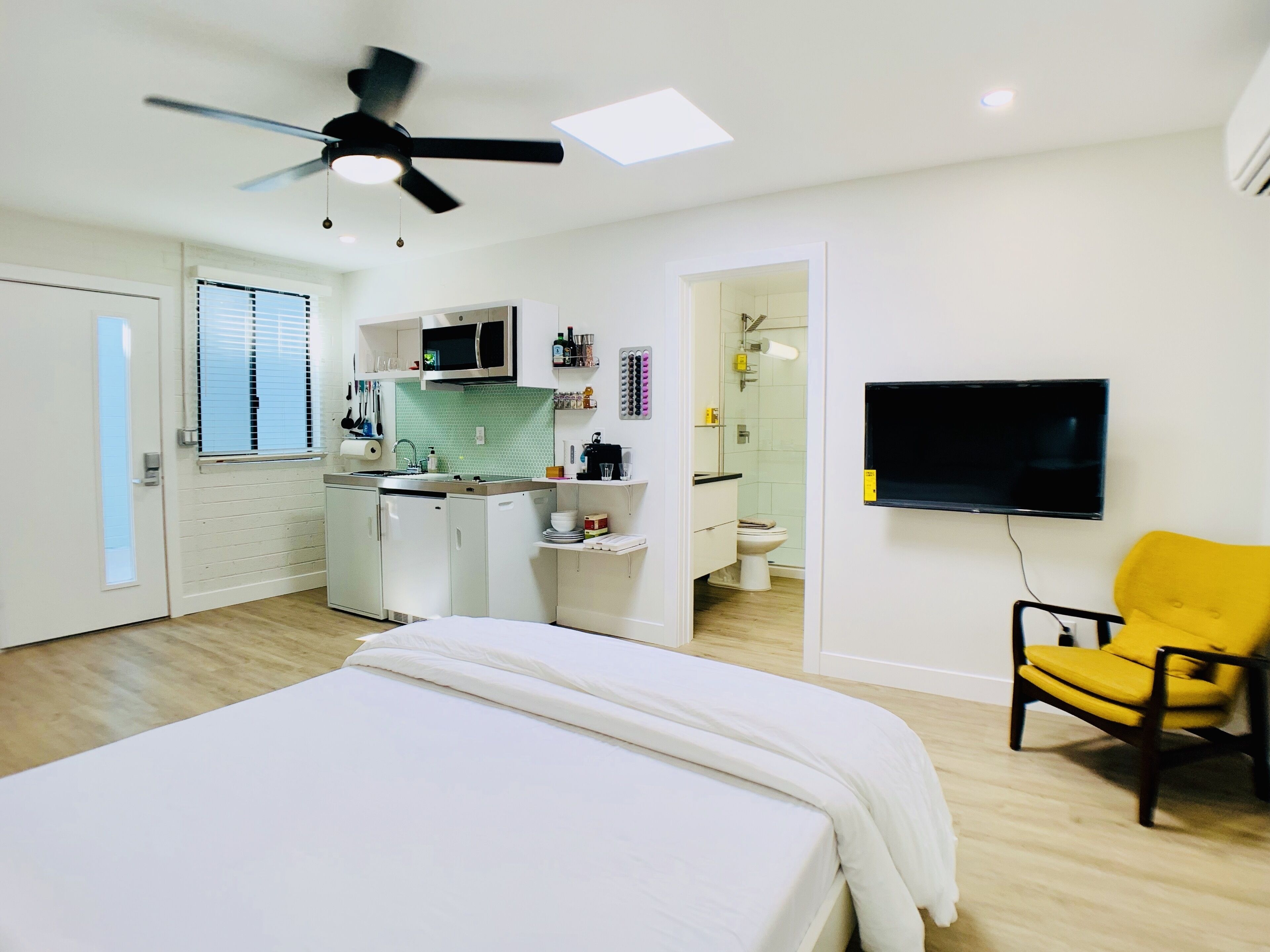 A modern hotel room with a bed, a yellow armchair, a TV, a kitchenette, and a bathroom.