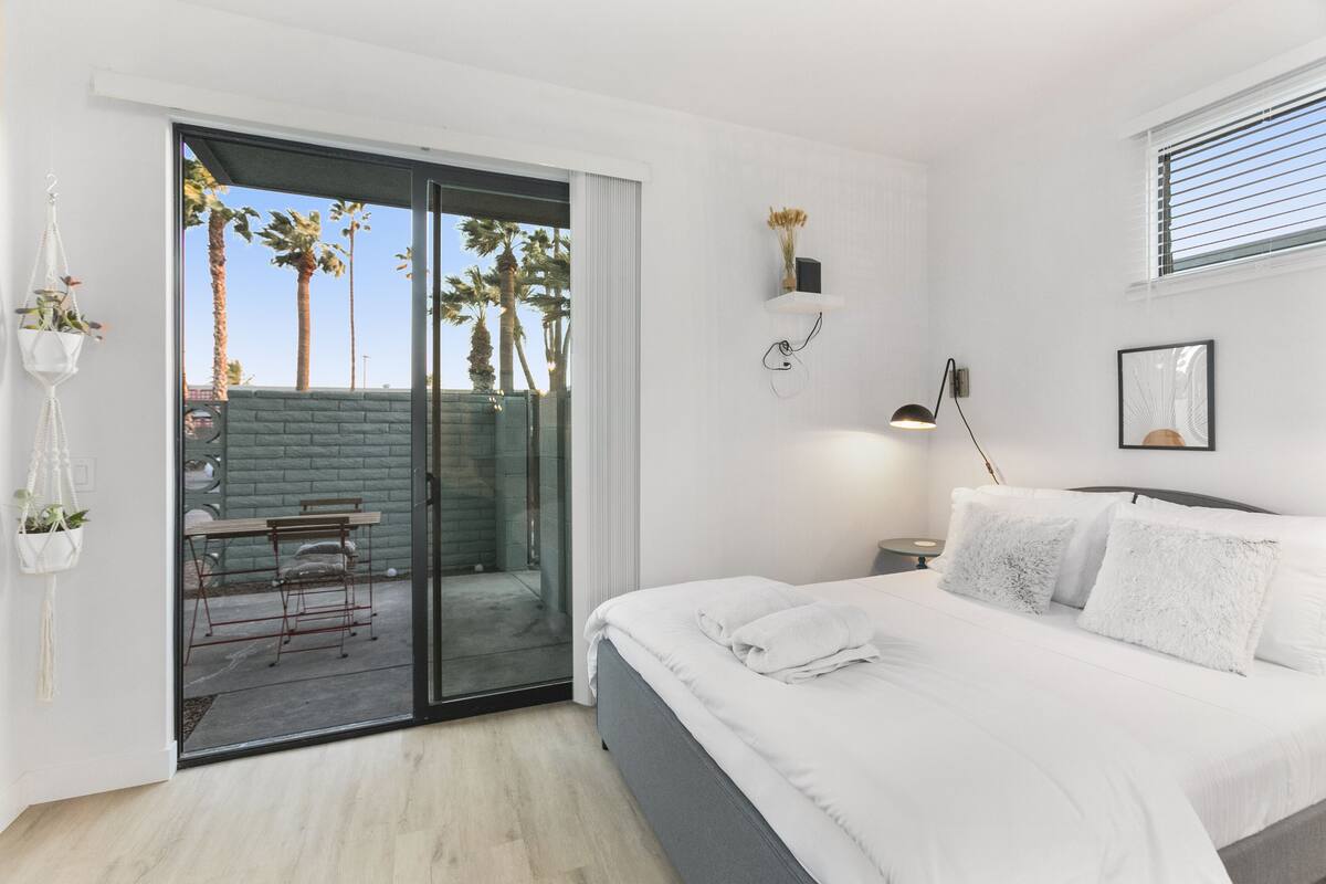A bedroom with a bed, a nightstand, a wall-mounted lamp, a framed picture, and a sliding glass door leading to an outdoor area with a table and chairs.