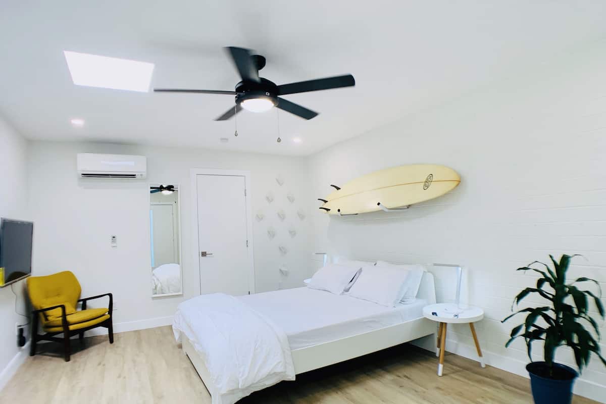 A bedroom with a bed, a ceiling fan, a surfboard, a chair, a small table, and a potted plant.