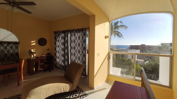 A room with a bed, a desk, a chair, a fan, a window with a view of buildings and palm trees, and a ceiling fan. - 2 br 2 ba in gated community w/private beach (Cabo San Lucas)