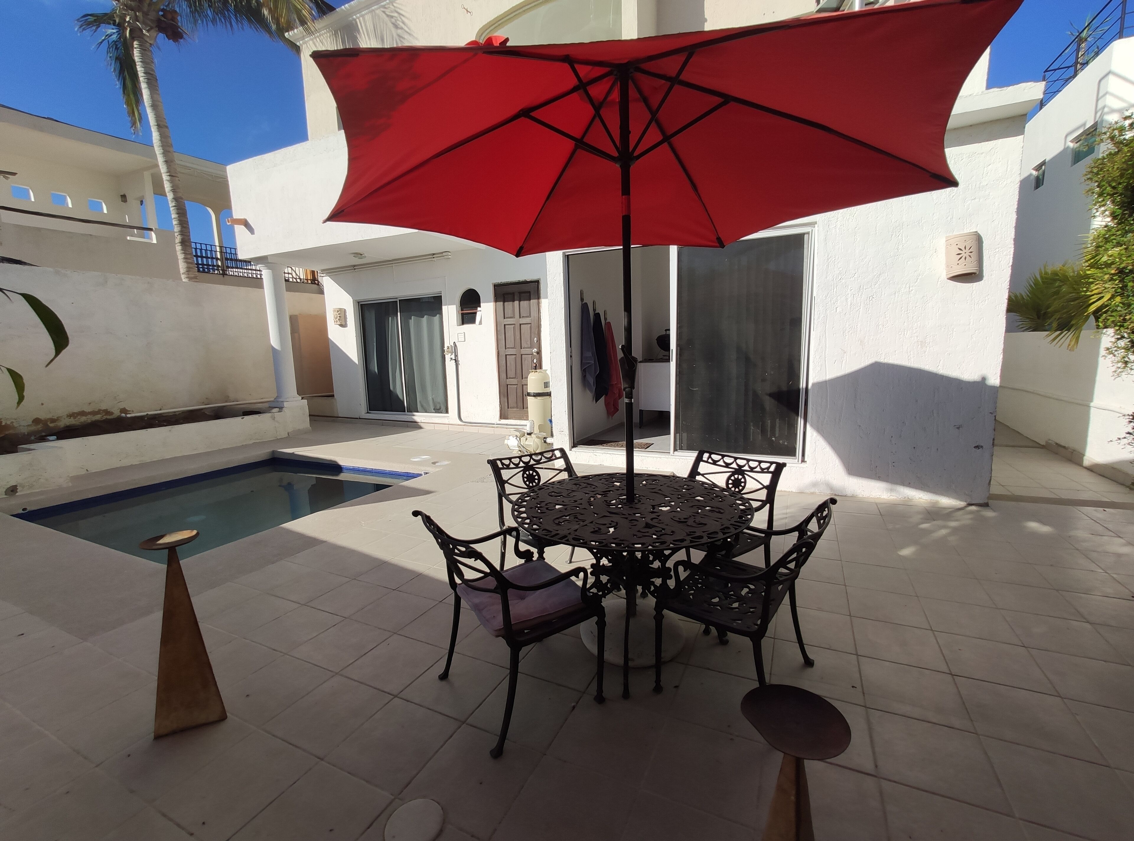 A poolside area with a round table and chairs, a red umbrella, and a small stool.