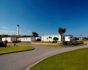 A caravan park with white mobile homes, a central walkway, and a lighthouse in the background. - Taid's Retreat Silversands Cove Beach Lossiemouth (Lossiemouth)