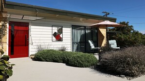 A single-story house with a red door, a covered porch with a chair, and a large window.