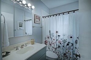 A bathroom with a white sink, two faucets, a mirror with lights, a shower curtain with a floral pattern, and a toilet. - 7 minutes from Providence Farm, MS Blues Trail and The Petrified Forest! (Flora)