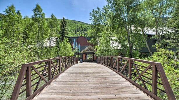 A wooden bridge leading to a lodge with a red roof, surrounded by lush greenery and a mountain in the background. - Bright Downtown Telluride Condo - Steps to Lift! (Telluride)