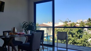 Ein Essbereich mit Tisch für zwei Personen, einer Kaffeekanne und einer Obstschale. Ein großes Fenster bietet Blick auf Stadt und Grünflächen.