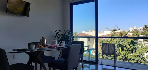 A dining area with a table set for two, a coffee pot, and a bowl of fruit. A large window offers a view of a cityscape and greenery.