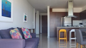 Ein modernes Wohnzimmer mit einer Couch, farbenfrohen Kissen, einem Essbereich mit Stühlen, einer Küche mit Wasserspender und Kunstwerken an den Wänden.