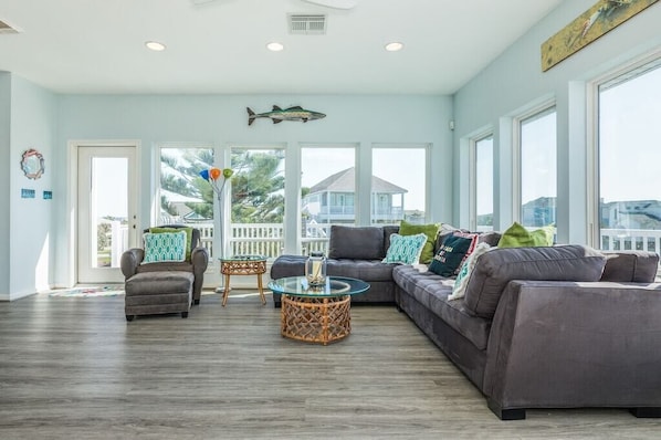 A spacious living room with a gray sofa, a glass coffee table, and a wicker side table. There are large windows offering a view of the outside.
