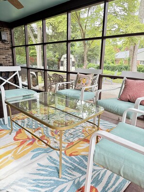 A sunroom with a glass table, white cushioned seating, and a view of the outdoors.