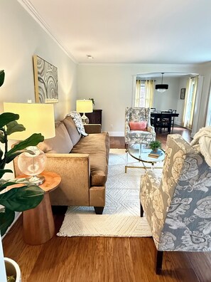 A living room with a sofa, armchair, coffee table, and a lamp. There is a dining area in the background with a table and chairs.