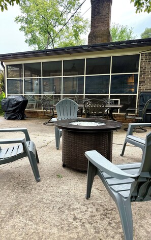 Een terras buiten met rieten meubels, een ronde tafel met vuurkorf, en een bakstenen gebouw met grote ramen op de achtergrond.
