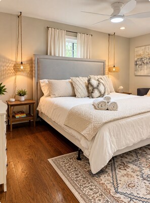 A bedroom with a large bed, a ceiling fan, a window with curtains, a bedside table with a lamp, and a rug.