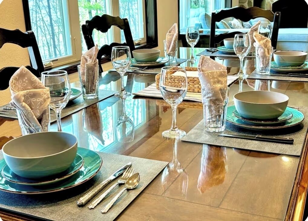Une table à manger est dressée avec de nombreux bols, verres et couverts, disposés sur une table en bois autour de laquelle sont placées des chaises.
