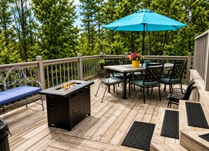 A wooden deck with a fire pit, outdoor table and chairs, a blue umbrella, and a potted plant.