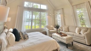 A spacious bedroom with a large bed, a sitting area with a sofa and armchair, a ceiling fan, and a view of the outdoors through large windows. - LAKEFRONT Mini-Resort Sleeps 32! Pool, hot tub, kayaks, fire pits, & more! (Cedar Bluff)