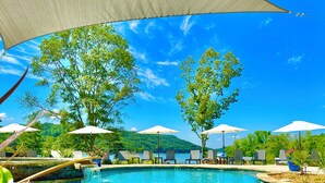 A pool area with clear blue water, surrounded by lounge chairs and umbrellas, and a view of a forested hill in the background. - LAKEFRONT Mini-Resort Sleeps 32! Pool, hot tub, kayaks, fire pits, & more! (Cedar Bluff)