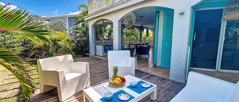 A covered outdoor patio with white furniture and a dining table set for two, surrounded by tropical plants.