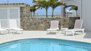 A poolside area with white lounge chairs, a stone wall, and a view of the sea. - Princess Anaelle · Beachfront, Lage Pool, Privacy, Orient Bay, Deluxe 2 BDR (SAINT MARTIN)