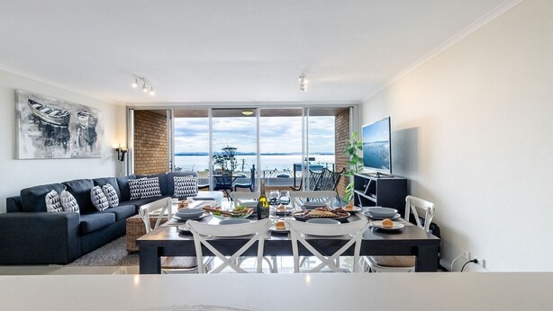 Una sala de estar moderna con comedor, ventanas amplias con vista al mar, televisor de pantalla plana y un sofá confortable.