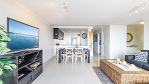 Una sala moderna con televisor de pantalla plana, comedor con sillas blancas, cocina con gabinetes blancos y sofá de mimbre con cojín estampado.