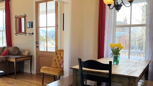 A cozy living room with a wooden table, a tufted armchair, a sofa, and a window with red curtains.