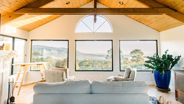A spacious living room with a high ceiling, large windows offering a view of a bridge and greenery, and a cozy seating area. - PRIVATE Hilltop Home: Stunning Carmel Views, Hot Tub, 3 Balconies, Espresso (Carmel-By-The-Sea)