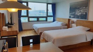 Habitación de hotel con dos camas, un escritorio, una silla, una lámpara y un amplio ventanal con vistas a la ciudad.