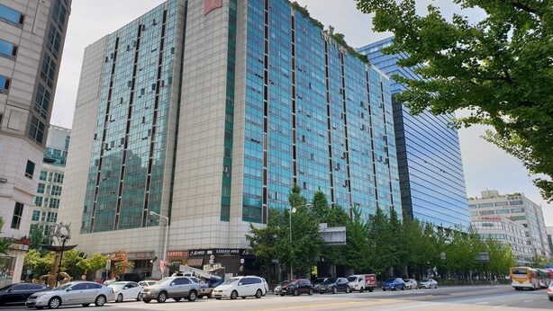 Un edificio moderno de varios pisos con fachada de vidrio, rodeado de automóviles estacionados y árboles.