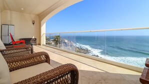Uma varanda com móveis de vime, guarda-corpo de vidro e vista para o mar.