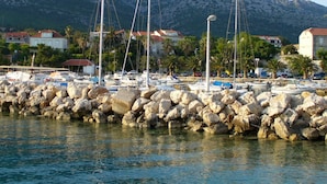 A marina with boats docked, rocky breakwaters, and houses in the background with mountains.