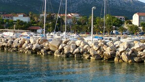Un puerto deportivo con barcos amarrados, rompeolas rocosas y casas al fondo, con montañas.
