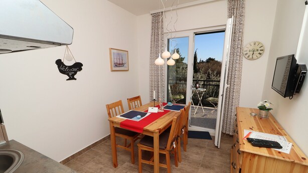 A dining area with a wooden table, chairs, and a red placemat. There is a window with curtains, a clock on the wall, and a television on a wooden cabinet. - Apartments Trstenica-One Bedroom Apartment with Balcony (Orebic)