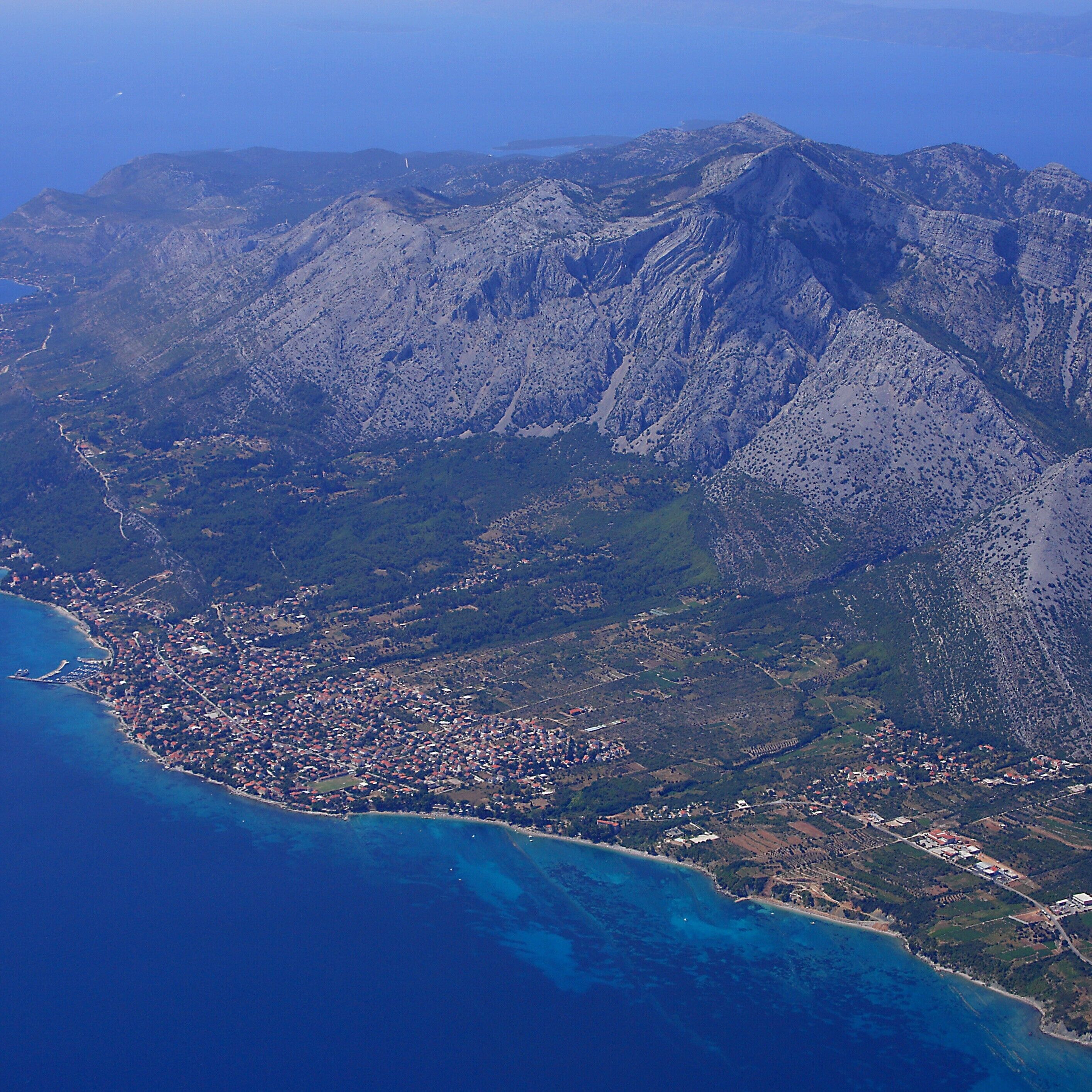 A coastal town with a mountainous backdrop, featuring a mix of residential and commercial buildings, a coastline, and a clear view of the surrounding terrain.