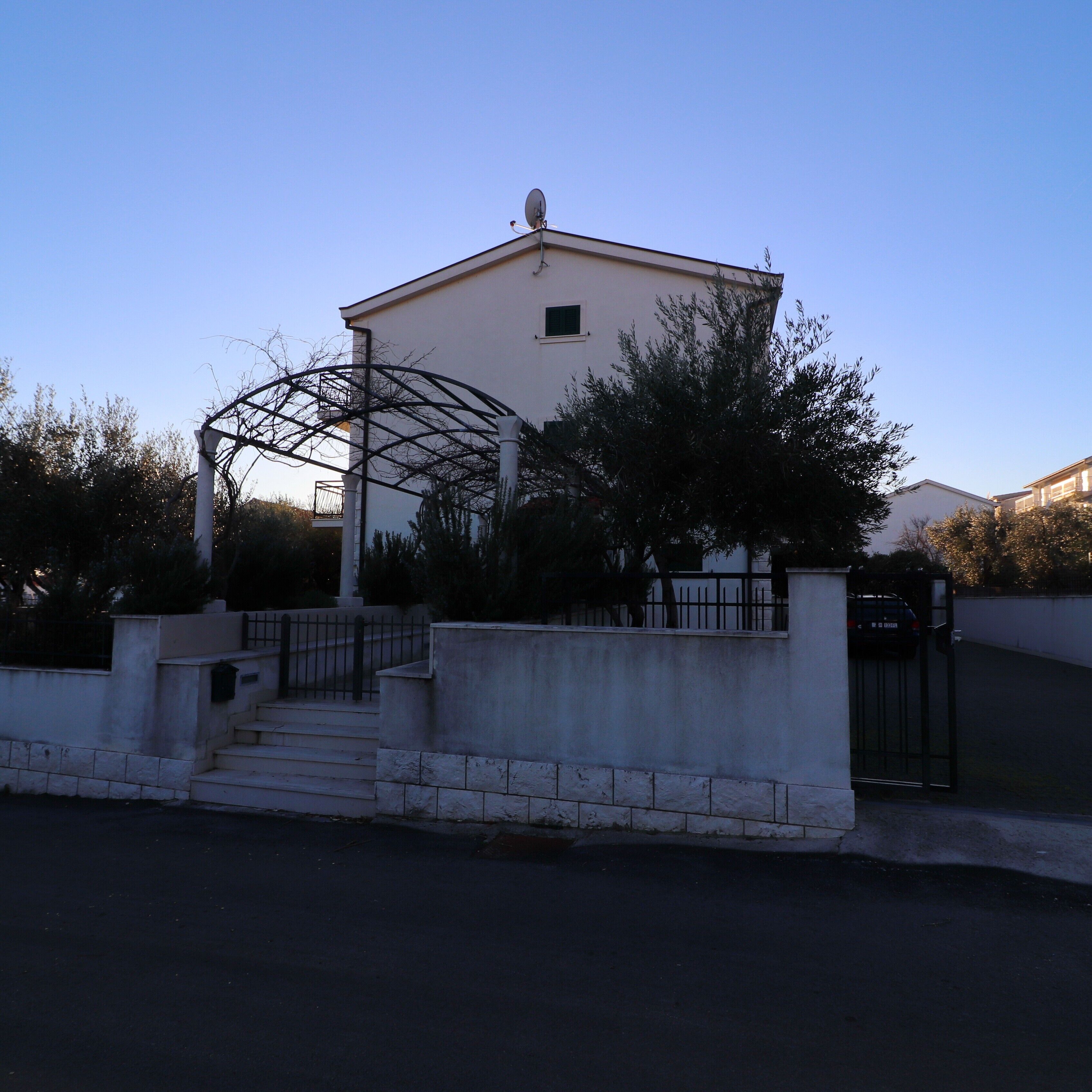 Un bâtiment à l’extérieur blanc, avec une petite fenêtre et une antenne parabolique sur le toit. On y trouve une porte en métal et un escalier menant à l’entrée.