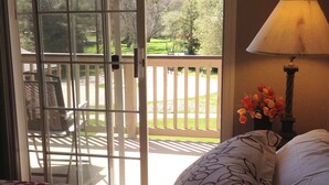 Une chambre avec un lit, une table de chevet avec une lampe, et une vue sur un balcon aménagé avec du mobilier d’extérieur et un jardin, accessible par une porte-fenêtre coulissante.