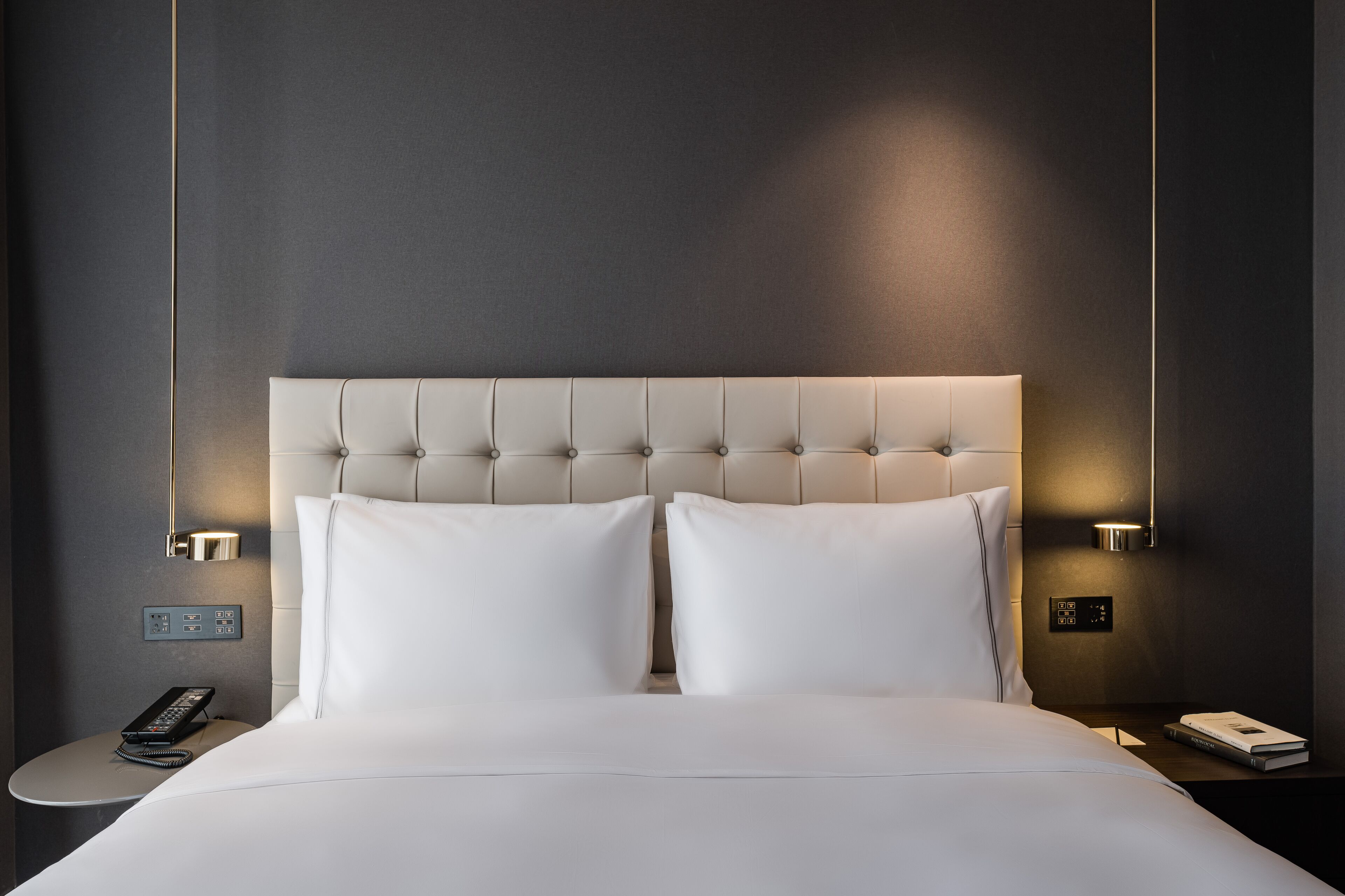 Une chambre d’hôtel moderne avec une tête de lit capitonnée, deux oreillers blancs, une table de chevet avec un téléphone, une lampe et un livre.