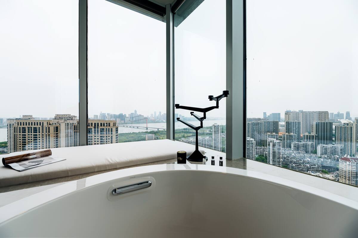 Un baño moderno con una bañera grande, un ventanal con vistas a la ciudad y una mesita con una revista y una botella.