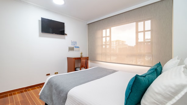 Ein Hotelzimmer mit einem Bett, einem Schreibtisch, einem Sessel, einem an der Wand befestigten Fernseher und einem Fenster mit Jalousien.