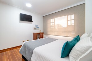 Ein Hotelzimmer mit einem Bett, einem Schreibtisch, einem Sessel, einem an der Wand befestigten Fernseher und einem Fenster mit Jalousien.