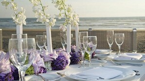 A table set for a meal with white tablecloths, purple and white flowers, and clear glassware against a beach backdrop. - Mayan Palace In Acapulco inside Vidanta Resort Luxury in your services (Acapulco)