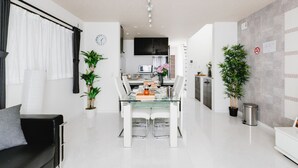 A modern living room with a glass dining table, white sofas, and potted plants.