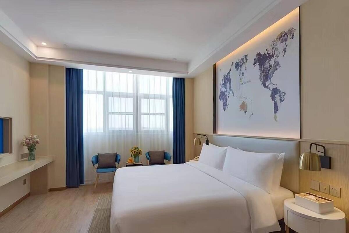 A hotel room with a large bed, a desk with a chair, a world map on the wall, and a window with curtains.