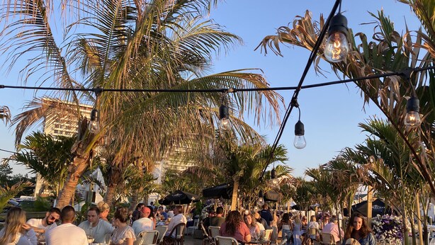 An outdoor dining area with wooden deck chairs and tables, surrounded by palm trees and under string lights.